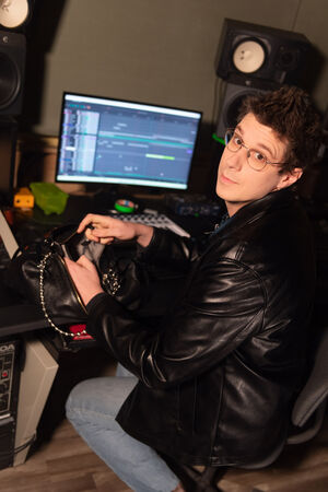 A person wearing glasses and a black leather jacket by INA KENT sits at a desk with a computer screen displaying audio editing software, reaching into a black bag.
