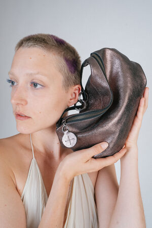 Person with short hair holds a metallic INA KENT zipper bag close to their face, looking to the side.