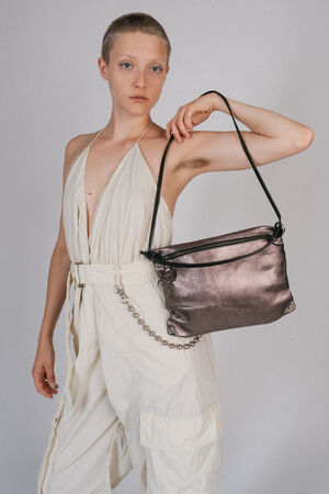 A person with short hair, dressed in a white jumpsuit, holds an INA KENT metallic shoulder bag. The bag features a black strap and chic chain detail. They stand against a plain background.
