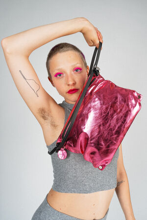 Person with short hair, bright pink eyeshadow, and a visible tattoo under their arm, holding a shiny pink INA KENT bag over their shoulder while wearing a gray sleeveless top.