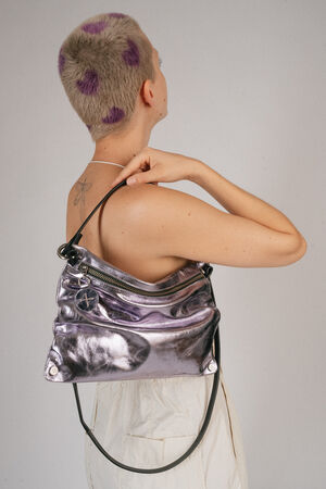 A person with short hair and purple spots holds an INA KENT shiny silver shoulder bag with black straps behind their back. They are looking over their shoulder, with a tattoo visible on their upper back.