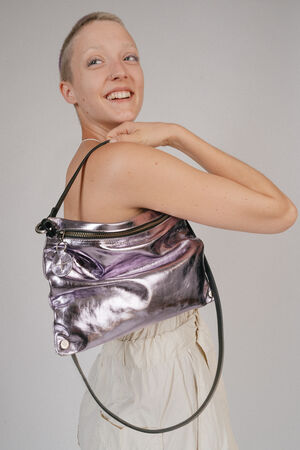 A person with a shaved head smiles while holding an INA KENT shiny metallic handbag over their shoulder. They wear a light-colored top and pants.