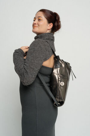 A person in a grey turtleneck sweater and dress stands facing sideways, looking back over their shoulder, carrying an INA KENT metallic backpack with a circular charm attached.