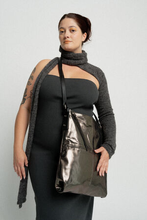 Eine Frau in einem dunklen trägerlosen Kleid und einem grauen Schal hält lässig eine metallische Tasche von INA KENT. Sie hat ein Tattoo auf ihrem linken Arm und steht vor einem schlichten weißen Hintergrund.