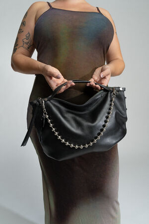 A person in a sleeveless, multicolored dress is holding an INA KENT black leather handbag with a chain detail. The person's arms have visible tattoos.