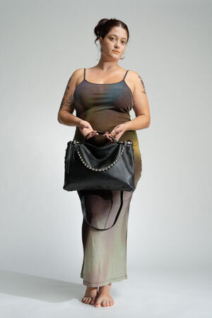 Woman stands barefoot on a plain backdrop, holding an INA KENT black bag with a beaded chain strap. She wears a sleeveless, multicolored dress and has tattoos on her arms.