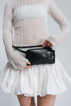 A person wearing a semi-sheer white long-sleeve top and a white skirt, holding an INA KENT small black handbag with a belt around their waist.