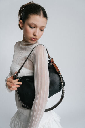 A woman in a white, semi-sheer long-sleeve top holds an INA KENT black leather handbag over her shoulder, looking down.