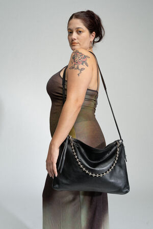 A person stands facing slightly left, wearing a sleeveless dress and holding an INA KENT black handbag with chain detail over their shoulder. They have a visible arm tattoo and are against a plain background.