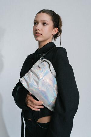 A person with tied-back hair in a black outfit holds an INA KENT shiny, iridescent bag against a plain background.