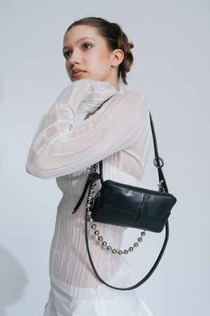 A woman with tied-back hair, wearing a white semi-transparent blouse and holding an INA KENT black rectangular purse with a chain strap, poses against a plain background.