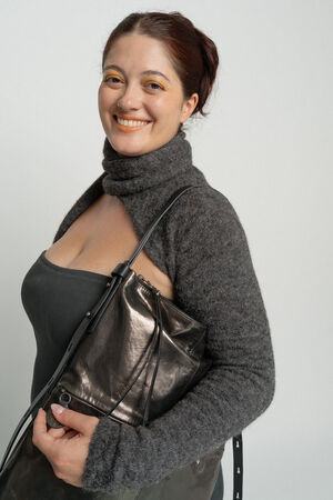 Eine Frau mit zurückgebundenem dunklem Haar lächelt und hält eine metallische INA KENT-Handtasche in der Hand. Sie trägt einen grauen Pullover über einem schwarzen Oberteil und steht vor einem schlichten hellen Hintergrund.