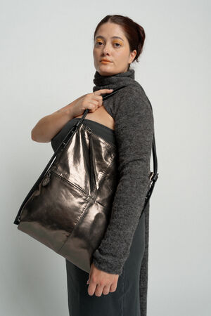 A person with a neutral expression holds a large metallic INA KENT bag while wearing a grey, long-sleeved turtleneck sweater and dark pants.