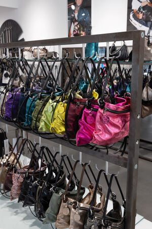 Eine Ausstellung verschiedenfarbiger Handtaschen, darunter violette, grüne, gelbe, schwarze und rosafarbene Modelle von INA KENT, ordentlich angeordnet auf Metallregalen in einem gut beleuchteten Laden.