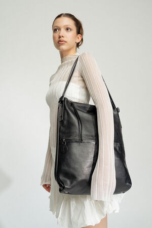 Eine Person in einem weißen Kleid und einem transparenten Langarmoberteil hält vor einem schlichten Hintergrund eine elegante schwarze Lederhandtasche von INA KENT über der Schulter.
