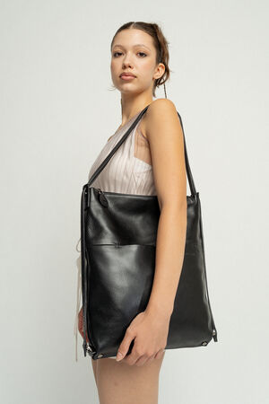 A person with a neutral expression carries a large black leather INA KENT tote bag over their shoulder, wearing a sleeveless light-colored dress against a plain white background.