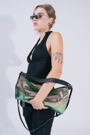 A person wearing sunglasses, a sleeveless black top, and black pants holds a large iridescent INA KENT handbag. The person has a tattoo of an animal on their left upper arm.