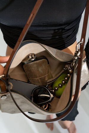 Eine Handtasche von INA KENT gibt ihren Inhalt preis: eine Brieftasche, ein grüner Apfel, eine Brille und ein Smartphone, alles gehalten von einer Person in einem schicken schwarzen Oberteil.