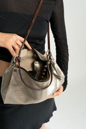 Eine Person im schwarzen Outfit hält eine geöffnete metallische INA KENT-Handtasche in der Hand und gibt den Blick frei auf den Inhalt, darunter ein Telefon und einen mit Perlen besetzten Schlüsselanhänger.