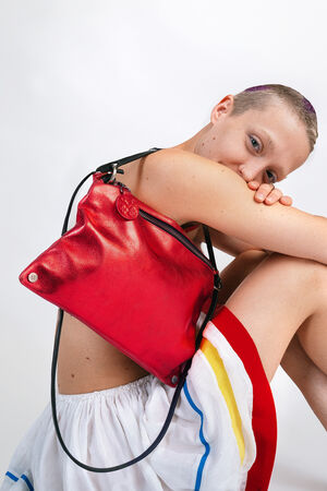 Person with short hair holding an INA KENT red leather handbag, sitting with knees up, wearing a white dress with colorful stripes.