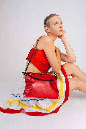 Person in a red top and colorful skirt seated with an INA KENT red handbag over their shoulder, gazing upwards.