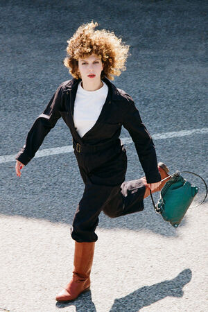 A person with curly hair in a stylish INA KENT jumpsuit and red boots is running on a concrete surface, holding a vibrant green bag.