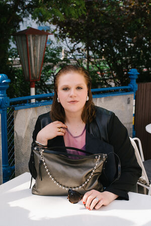 A person with wet hair sits at an outdoor table, holding an INA KENT black bag with a pearl strap and sunglasses attached. Trees and a blue fence are in the background.