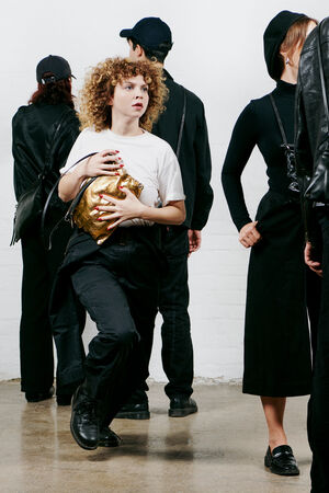 A woman with curly hair holding a shiny gold INA KENT bag stands among a group of people dressed in black, who are facing away.