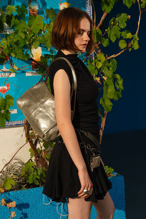 A person with short brown hair wearing a black dress and a metallic leather flap bag by INA KENT. The person stands outdoors near a blue wall covered with green vine leaves.