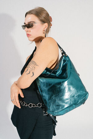 Eine Person steht vor einem weißen Hintergrund und trägt ein elegantes schwarzes Outfit und eine Sonnenbrille. Sie trägt eine metallisch blaugrüne Tasche von INA KENT über der Schulter und zeigt ein auffälliges Hirschtattoo auf ihrem Arm.