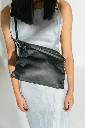 A close-up of the MOONLIT ed.1 shoulder bag made from natural black leather. The bag is worn crossbody and emphasizes its versatility and modern silhouette in combination with a silver-colored outfit.