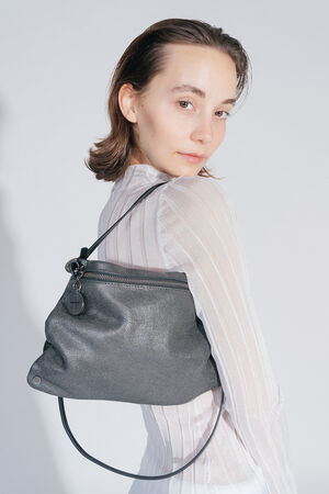 Woman in a white, translucent top and white skirt holds the INA KENT bag MOONLIT ed.1 in her hand. The bag has a textured, black surface and is presented close to the upper body.