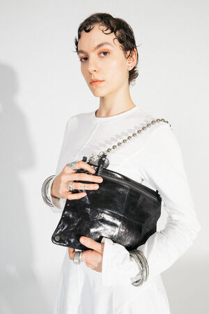 BALL'N'CHAIN metallic deep tar (worn, close to the shoulder): A shiny silver ball chain used as a shoulder strap for a black bag. The necklace and bag are presented by a person in a white outfit.