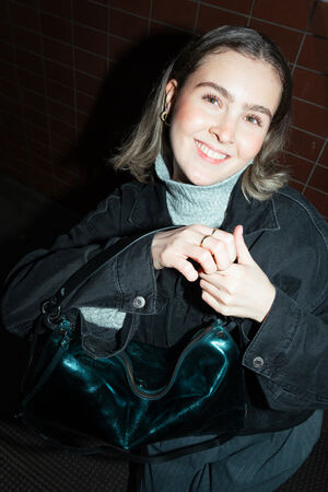 A female-presenting person smiles into the camera while holding a shiny blue-green bag by INA KENT named DINKUM ed.2. She holds the bag with both hands.