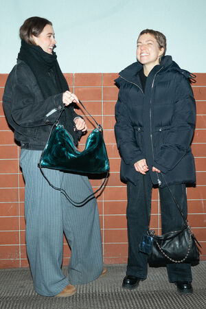 Two female-presenting people stand laughing in front of an orange-brown tiled wall. The person on the left is holding a large, shiny bag in dark green (“DINKUM ed.2” model in crackled dark teal leather), while the person on the right is carrying a smaller black ORNAMENT ed.2 bag with silver-colored details and a small blue X.LOMI ed.2 mini wallet.