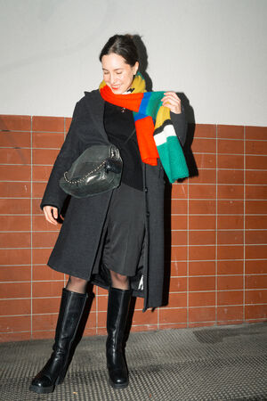 A female-presenting person stands frontally, wearing a black sweater, a colorful striped scarf and black boots. The bag “EILEEN ed.1” in crackled graphite hangs over her shoulder.