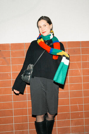 A female-presenting person is wearing a black coat, black boots and a colorful striped scarf. She is holding the “EILEEN ed.1” bag in crackled graphite over her shoulder.