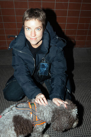 A female-presenting person in a dark jacket is kneeling next to a dog with a colorful harness. She is carrying a black bag from INA KENT with a mini wallet (X.LOMI ed.2) in blue metallic. 