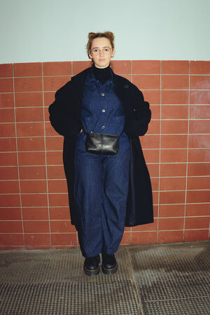 A female figure in a black coat and a dark blue two-piece suit looks directly into the camera. She stands in front of a wall with red tiles and carries an INA KENT bag “X.MOG” made of black leather around her waist. 