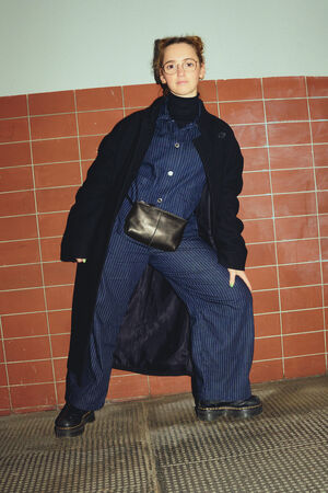 A female figure in a black coat and a dark blue two-piece suit looks directly into the camera. She is carrying an INA KENT bag “X.MOG” made of black leather around her waist.