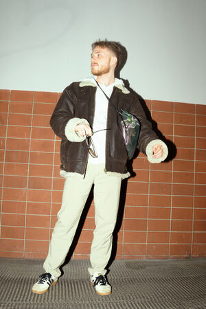 A male-presenting person stands frontally in front of a tiled wall and looks to the side with a smile. He is wearing a dark brown leather jacket with a light lining, a white hooded jacket, light-colored pants and black and white sneakers. Over his shoulder, he is carrying a shimmering, multi-colored bag (“DINKUM ed.2”) in a dazzling design.