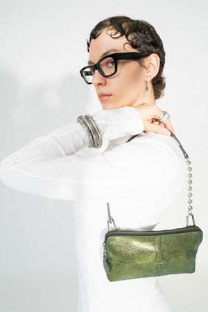 A female model with short, wavy hair carries the INA KENT IVI ed.3 bag in crackled guacamole metallic leather. The small bag with silver ball chain and green sheen is the focal point, while the person wears a white, simple outfit and eye-catching accessories.