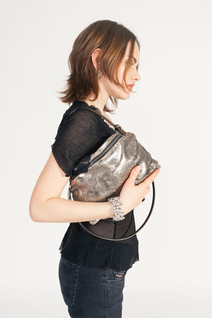 Female model in a black, transparent top and jeans holds the INA KENT MOONLIT ed.1 bag in metallic bright anthra close to her body. The bag has a shiny surface and a simple leather strap.