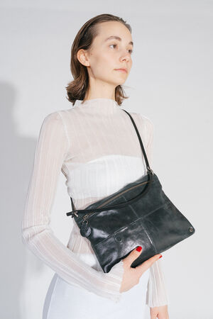 One person holds the MOONSTRUCK ed.2 bag made of crackled-tar-colored leather with one hand. The bag is worn cross-body, has a discreet black strap and a minimalist design. The person is wearing an elegant white outfit with red nails.