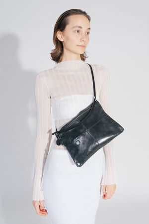 A person carries the MOONSTRUCK ed.2 bag made of crackled tar-colored leather across the body. The bag has a shimmering effect, visible zipper and a minimalist black strap. The person wears a transparent white top and looks to the side.