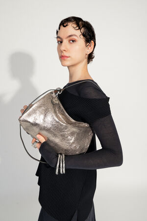 Model with INA KENT ORNAMENT ed.2 metallic bright anthra (front view, worn): A metallic anthracite-colored bag worn under the arm by the model. The bag has a shiny surface and a slim, brown shoulder strap. The model is wearing black clothing and accessories with futuristic details.