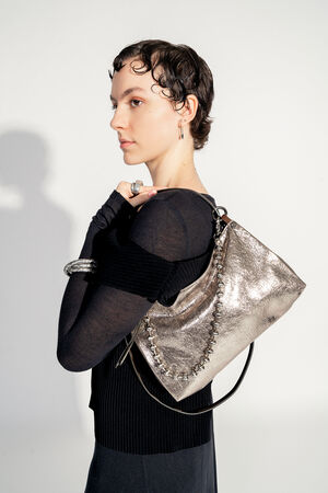 Model with INA KENT ORNAMENT ed.2 metallic bright anthra (side back view): The model elegantly carries the bag over her shoulder, focusing on the shiny surface and the decorative ball chain. The model turns slightly to the side to emphasize the details of the bag.