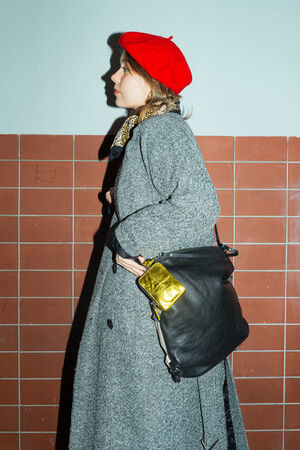 A female-presenting person stands in profile wearing a long, gray coat, a red beret and a black scarf with a pattern. A black bag by INA KENT (DELA EDEN ed.1) with a shiny gold-colored pendant hangs over her shoulder.