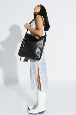 Eine weiblich gelesene Person trägt die Tasche AMPLE ed.1 in schwarzem Leder über der Schulter. Sie trägt ein silbernes, schimmerndes Kleid mit hohem Beinschlitz und weiße Stiefel. Die Person schaut leicht nach oben und das Bild ist minimalistisch mit einem weißen Hintergrund gestaltet.
