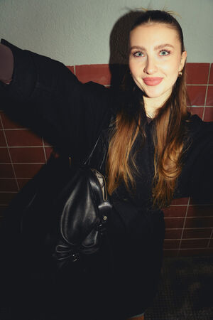 A female-presenting person is wearing a long black coat and smiling at the camera. She is carrying the “DELA EDEN ed.1” bag by INA KENT in pure black over her shoulder.